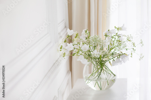 Bouquet of gentle bells in vase. Morning light in the room. Soft home decor, glass vase with white flowers on white wall background and on wooden table. Interior. Greeting card. Copy space.