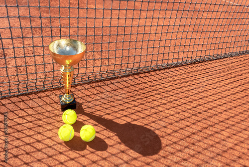 Trophy cup and tennis balls on a red clay court and shadow from the net. Champion or victory trophy. Winner in tennis tournament or championship concept. Sports background. Copy space © Elena