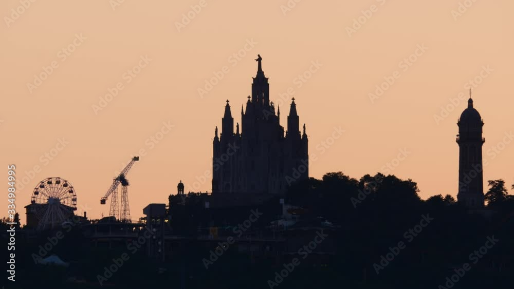 Silhouetted shot of Temple Expiatori del Sagrat Cor on top of Tibidabo hill, moving attractions of theme park seen around. Time lapse telephoto shot, shadow figure of Barcelona landmarks from distance