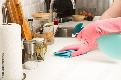 Manos con guante de látex rosa y botella de detergente pulverizante limpiando la encimera de la cocina. Vista de frente