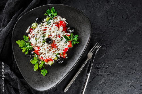 Healthy greek salad of fresh vegetables with tomatoes, lettuce, bell pepper, cucumbers, olives, feta cheese in plate on black stone background. Healthy food, vegetarian dieting, top view