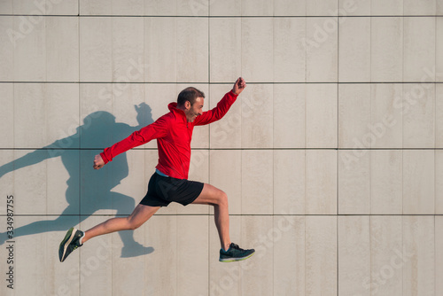 Caucasian athlete man in red jacket and black pants running and jumping on a sunny day with his shadow cast on a white wall.