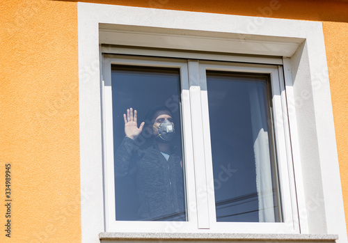 Coronavirus outbreak. Social distancing. Man in quarantine wearing protective mask looking through the window. Stay home. Spring outdoor