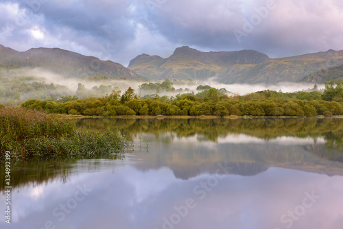 Peaceful Misty Morning With Perfect Reflections In Lake And Mountain Backdrop. Lake District, UK.