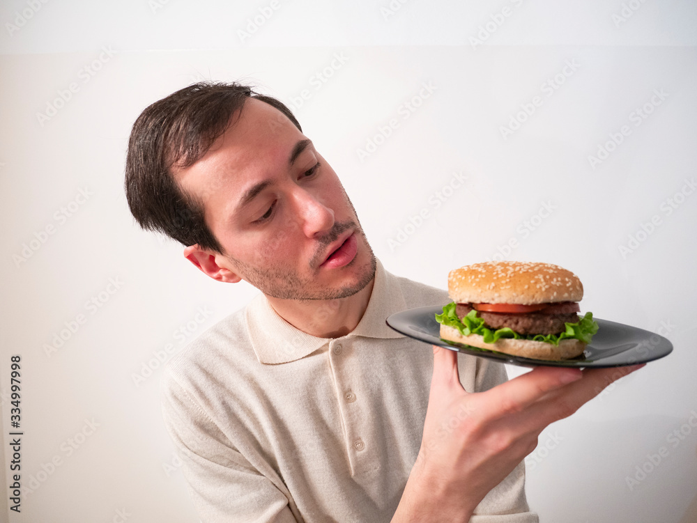 Handsome young man holding, looking and examining tasty hamburger for examination Handsome young man holding, looking and examining tasty hamburger for examination