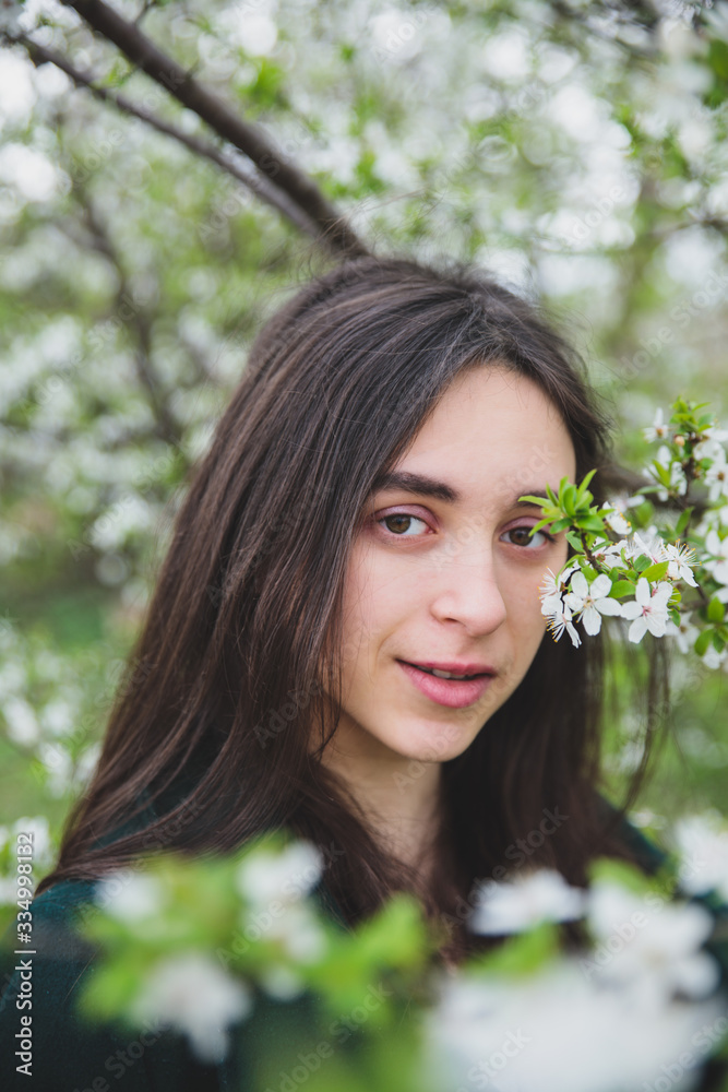 Young caucasian girl with dark hair in white t-shirt and green coat near beautiful white cherry blossom tree Young caucasian girl with dark hair in white t-shirt and green coat near beautiful white cherry blossom tree