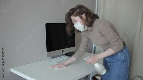 A young attractive girl in a quarantine protective mask thoroughly cleans the computer with a disinfectant.Coronavirus. COVID-19 photo