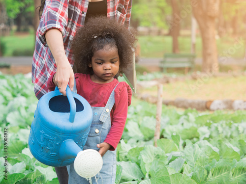 Cute adorable African girl using watering can water vegetable in garden with help from teacher or mother. Learning by doing, nature study for toddler.