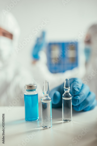 selective focus of biochemist in rubber gloves taking ampule near glass container with blue liquid