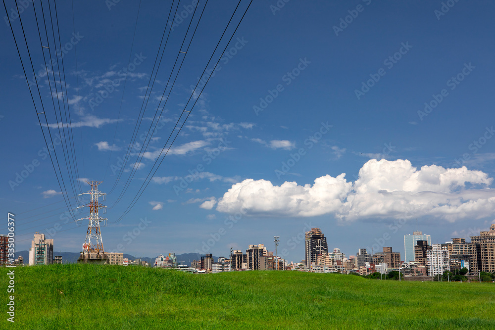 Electric tower on green grass in embankment park, city under blue sky and white clouds