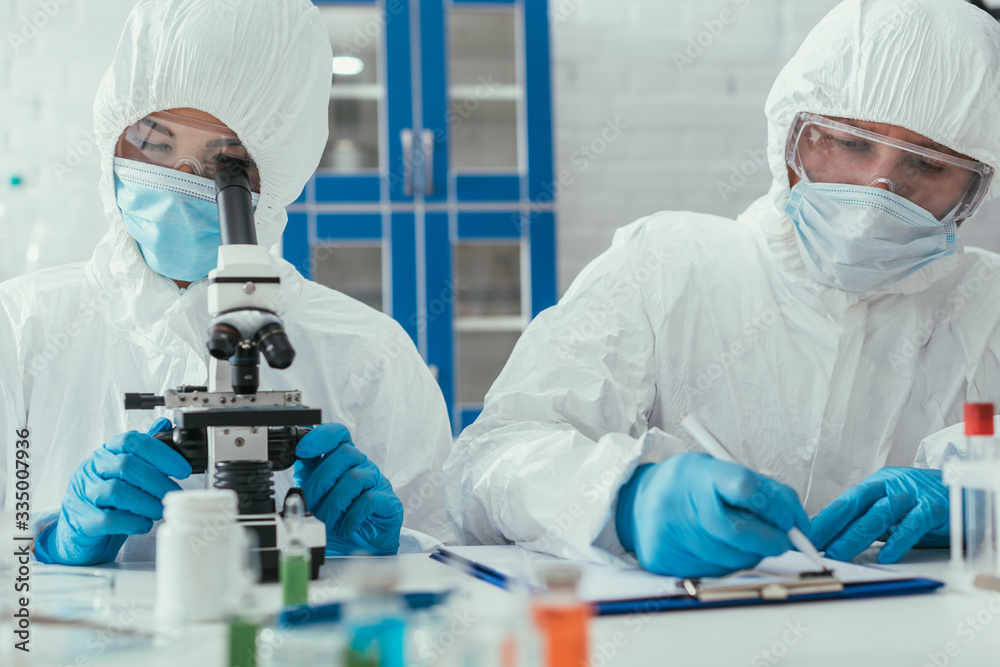 selective focus of biochemist looking through microscope near colleague writing down results on clipboard selective focus of biochemist looking through microscope near colleague writing down results on clipboard