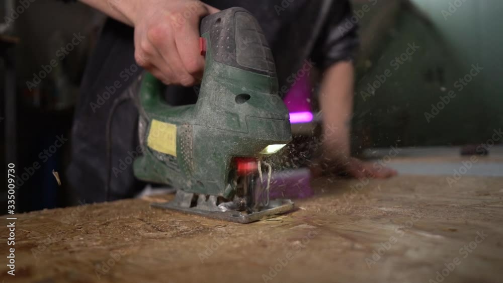Worker saws a wooden part with an electric jigsaw. Pink lamp in the background Worker saws a wooden part with an electric jigsaw. Pink lamp in the background