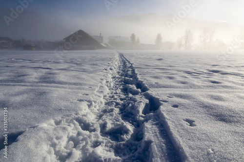 a winter landscape with a trodden out in snow pathway leading to countryside houses and mountains in the background that are shrouded in dense fog on a sunny morning in alps.