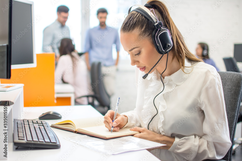 Young business woman working in front of computer, writing notes to notebook while sitting at her workplace in office Young business woman working in front of computer, writing notes to notebook while sitting at her workplace in office