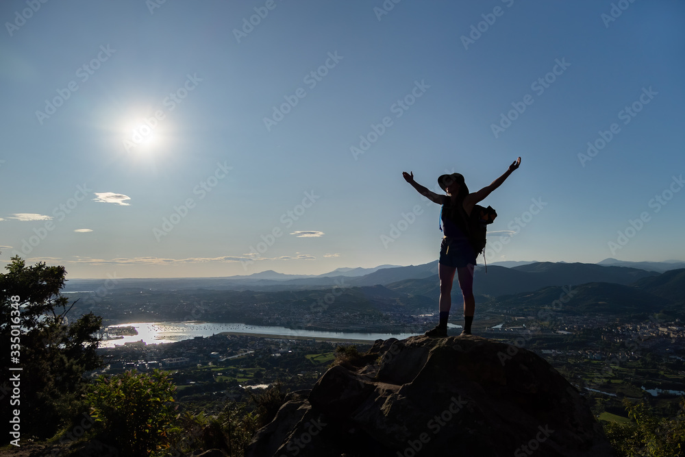 Silhouette of girl in hat, stains on a rock and spreads her hands to the sides. A city, mountains, sea and sun at the background Silhouette of girl in hat, stains on a rock and spreads her hands to the sides. A city, mountains, sea and sun at the background