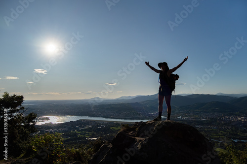 Silhouette of girl in hat, stains on a rock and spreads her hands to the sides. A city, mountains, sea and sun at the background