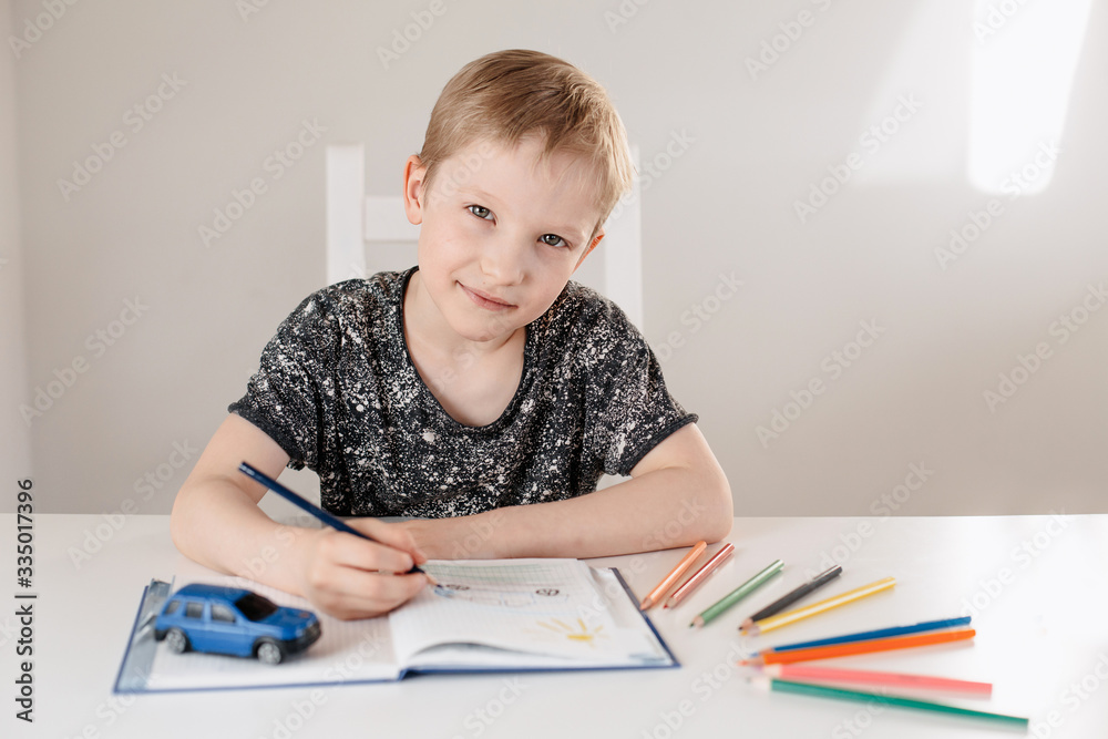 A child of European appearance is sitting at a table drawing with pencils in a white interior A child of European appearance is sitting at a table drawing with pencils in a white interior