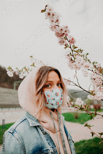 Attractive young woman in blue medical mask standing in the beautiful blooming tree  looking in the camera  urban background  sunny day. Natural light