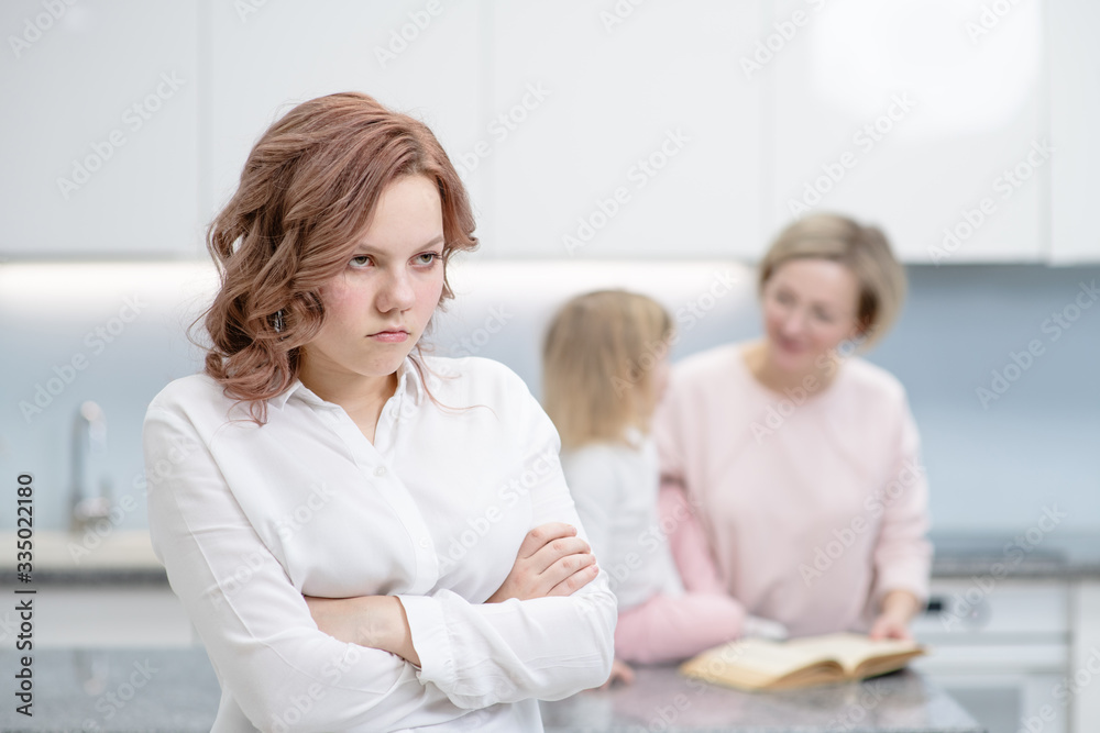 A teenage girl is standing with a sad expression on her face. Against the background, her mother plays with her younger sister. The problem of loneliness in the family concept
