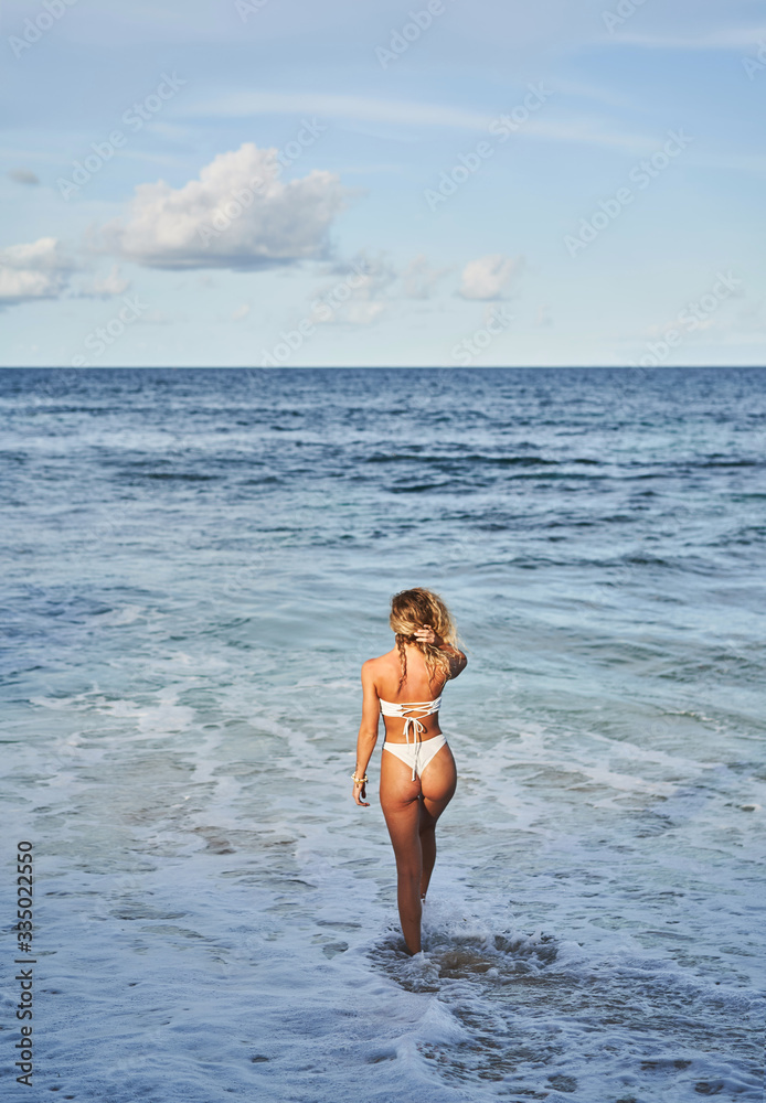 Slim woman in bikini carrying surfboard and walking on wet sand towards waving sea on beach