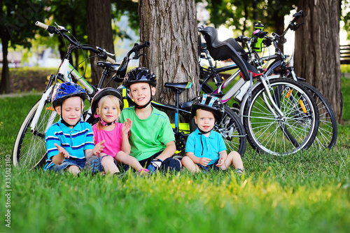a group of small happy preschool children in Bicycle safety helmets are smiling sitting on the fresh green grass in the Park against the background of bicycles and trees.