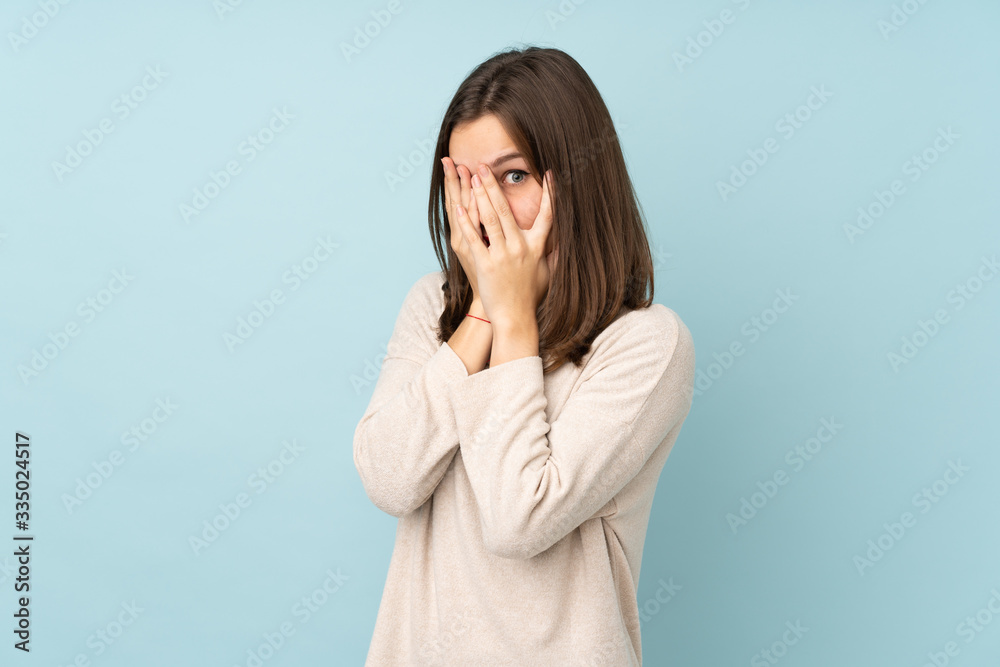 Teenager girl isolated on blue background covering eyes and looking through fingers Teenager girl isolated on blue background covering eyes and looking through fingers