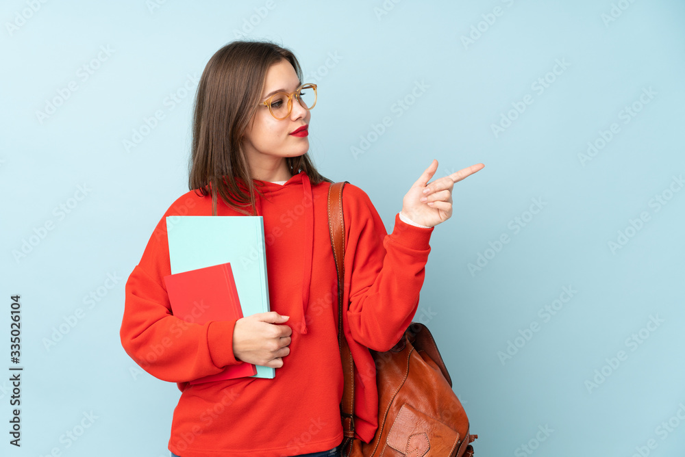 Student girl holding books isolated on blue background pointing finger to the side Student girl holding books isolated on blue background pointing finger to the side