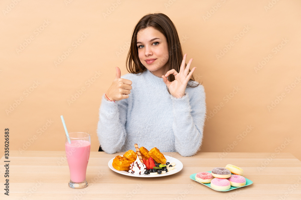 Teenager girl eating waffles isolated on beige background showing ok sign and thumb up gesture Teenager girl eating waffles isolated on beige background showing ok sign and thumb up gesture