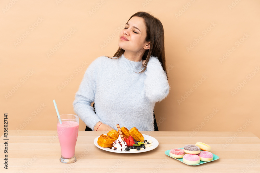 Teenager girl eating waffles isolated on beige background suffering from pain in shoulder for having made an effort Teenager girl eating waffles isolated on beige background suffering from pain in shoulder for having made an effort