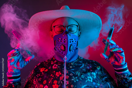 Strange man in medical respirator mask  glasses and hat holding two syrenges with coronavirus covid 19 test on red blue background.with smoke. Evil doctor  inventor of corona virus covid 19 conept