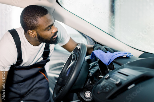 Concentrated professional African worker cleaning the car console and control panel with microfiber cloth. Car wash and detailing concept photo