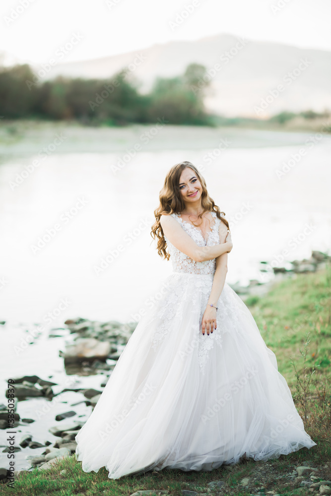 Beautiful brunette bride in elegant white dress posing near river