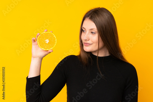Teenager caucasian girl holding an orange over isolated yellow background