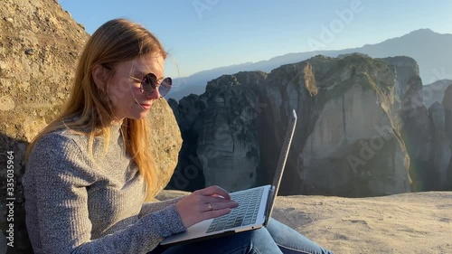 Gorgeous blonde woman-tourist at black glasses types text at laptop at the top of mountain with amazing view.