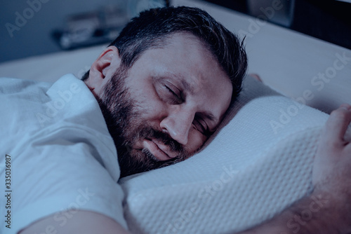 A lonely bearded dark-haired man sleeps soundly on his stomach at night on the bed. Close-up, orthopedic pillow, bedroom, self-isolation.