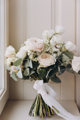 bride’s wedding bouquet of flowers and greenery with a ribbon stands near the window on a white background