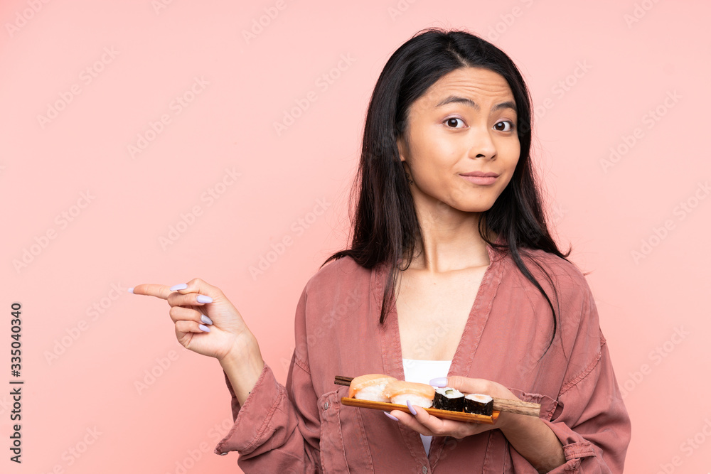 Teenager Asian girl eating sushi isolated on pink background pointing to the laterals having doubts Teenager Asian girl eating sushi isolated on pink background pointing to the laterals having doubts