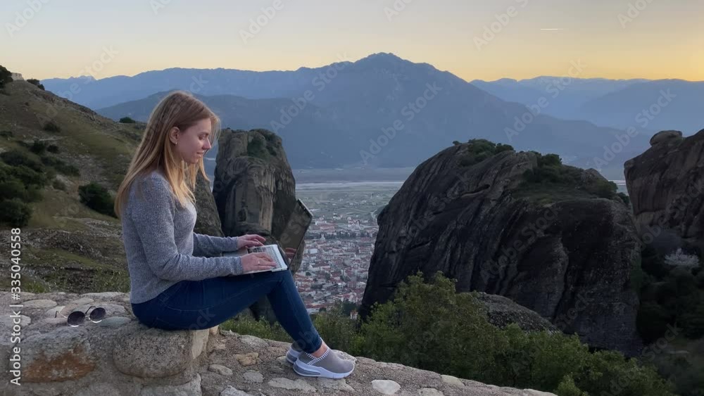 Young female tourist works remotely at laptop at Meteora mountains. Beautiful caucasian woman-freelancer works outdoor at computer in front of high rocks at sunset Young female tourist works remotely at laptop at Meteora mountains. Beautiful caucasian woman-freelancer works outdoor at computer in front of high rocks at sunset