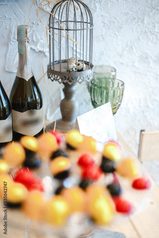 two bottles of champagne, Easter eggs in a decorative cage and glasses in the background, candies in colored glaze on a glass stand in the foreground out of focus