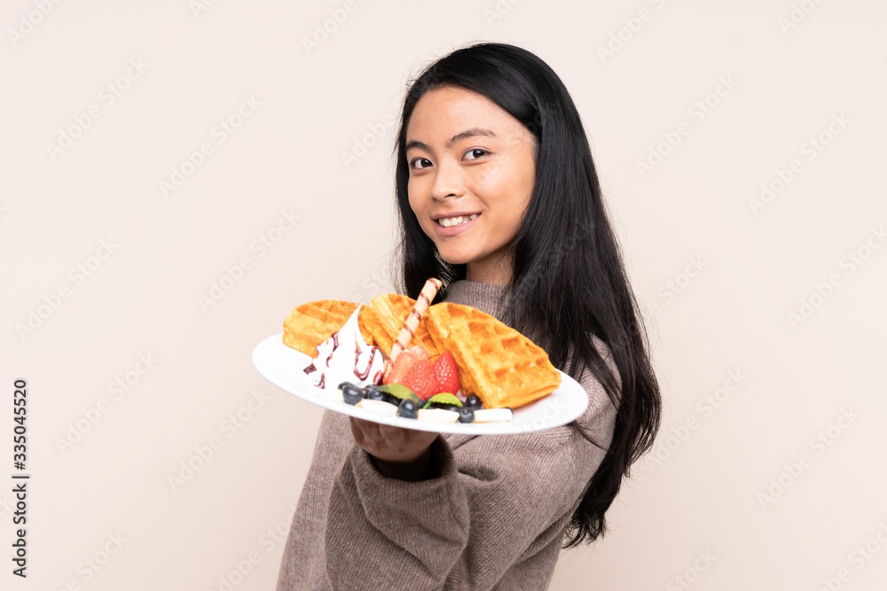 Teenager Asian girl holding waffles isolated on beige background with happy expression
