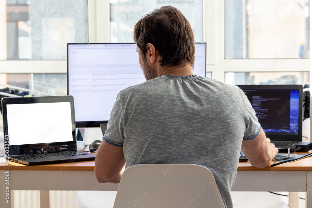 A man in a gray t-shirt is sitting at a workplace with two laptops and a monitor near the window. Remote work from home in quarantine. Back view - obrazy, fototapety, plakaty 