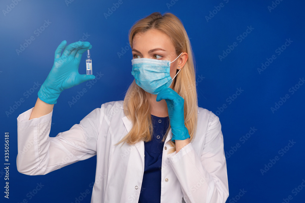 A girl doctor in a medical mask and gloves holds an ampoule with a vaccine against the disease on a blue background A girl doctor in a medical mask and gloves holds an ampoule with a vaccine against the disease on a blue background