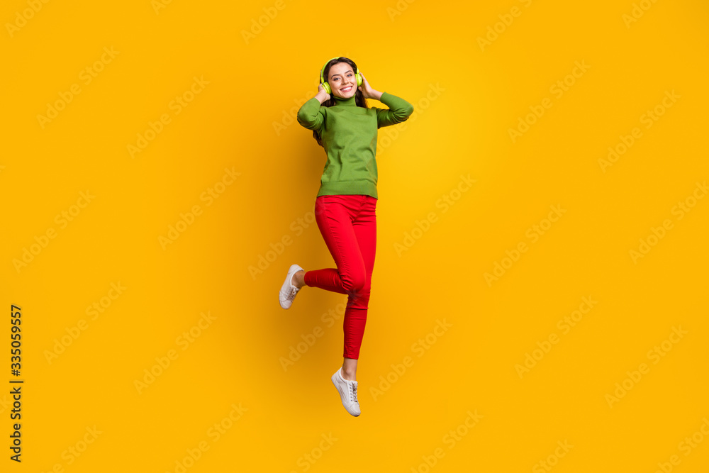 Full size photo of funky pretty lady jumping high holding ears with cool modern headphones listening music wear casual green jumper red pants isolated yellow color background