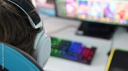 Locked down over the shoulder shot of a person wearing a gaming headset and using a backlit synchronising keyboard when playing a computer game