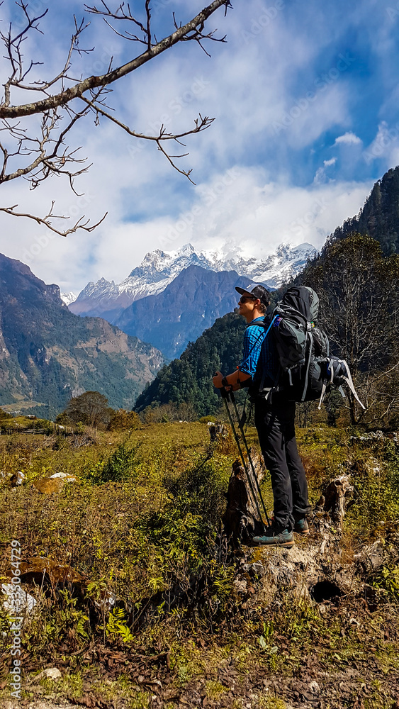 A man with a big black backpack admires Manaslu on his way on Annapurna Circuit Trek, Nepal. Forest to the right. To the left another Mountain. Manaslu covered with snow. Some flowers on the ground.