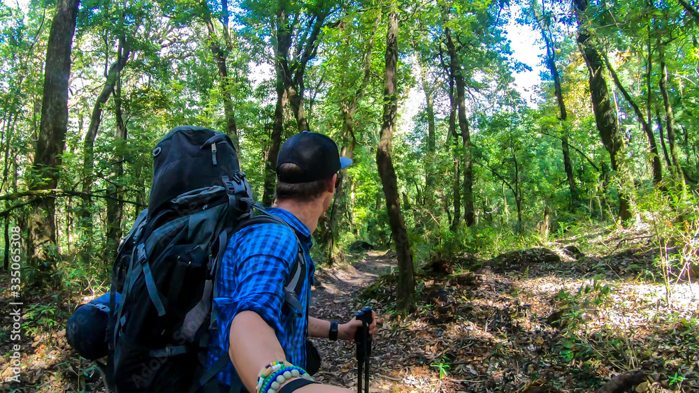 A man with a big black backpack hikes along a pathway in a forest on Annapurna Circuit Trek in Himalayas, Nepal, while taking a selfie. Dense trees. Exploration and adventure