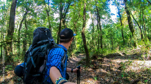 A man with a big black backpack hikes along a pathway in a forest on Annapurna Circuit Trek in Himalayas, Nepal, while taking a selfie. Dense trees. Exploration and adventure