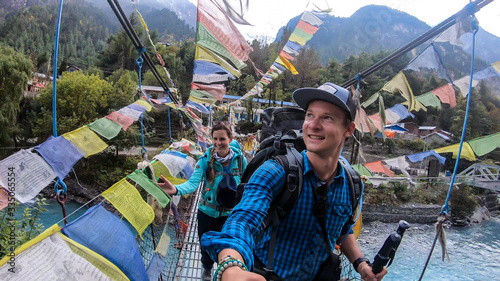 A couple in hiking outfits with big backpacks crossing an extension bridge on the Annapurna Circuit Trek in Himalayas, Nepal. There is a lot of colorful prayer flags attached to the bridge. Happiness