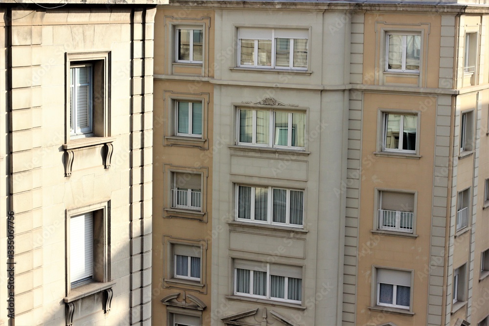sunny windows on the upper floors of two beige buildings, one in the foreground, the second in the background.
