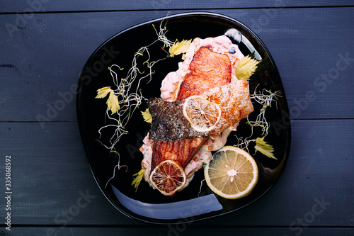 One piece of baked salmon grilled pepper, lemo, vegetable in white souse on a black plate. wood blue background